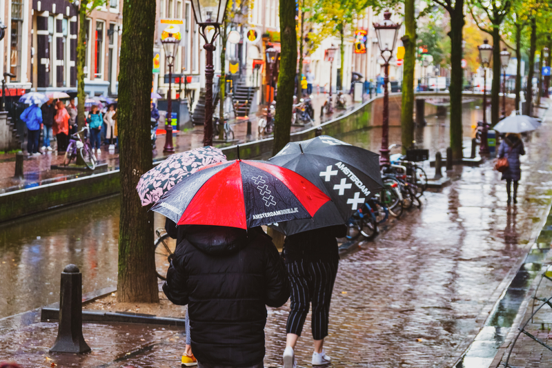 photo-of-two-people-walking-by-an-amsterdam-canal-while-holding-umbrellas