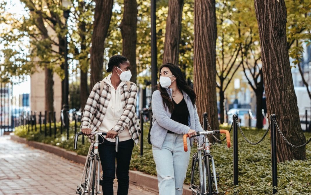 photo-of-two-girls-walking-with-bikes-and-face-masks