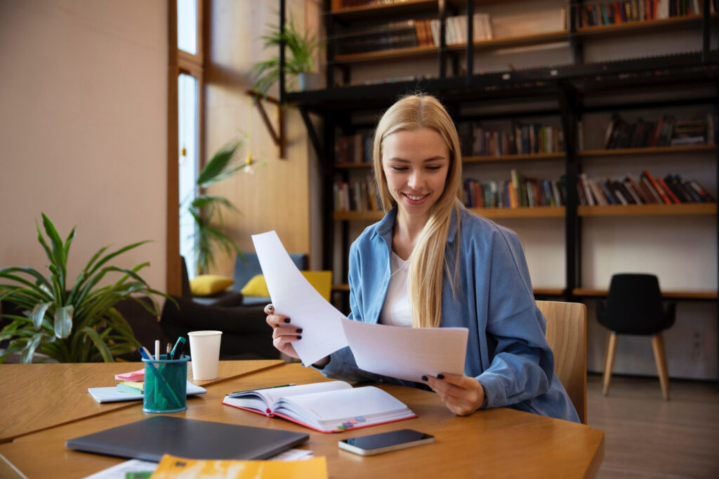 photo-of-young-woman-updating-resume-holding-sheets-of-paper-in-office-with-laptop-and-pens-on-table