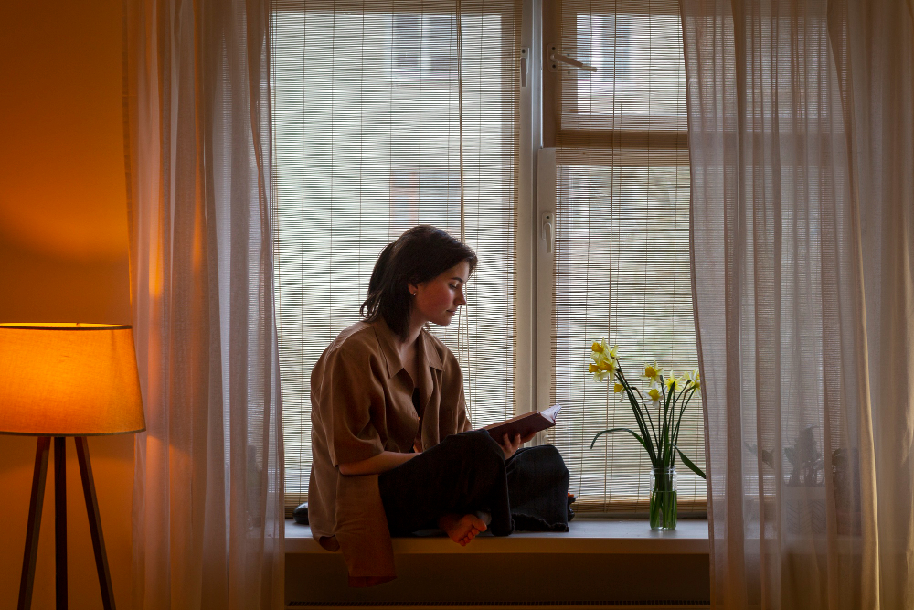 photo-of-woman-reading-in-windowsill-with-pretty-blinds-and-curtains-in-cosy-bedroom