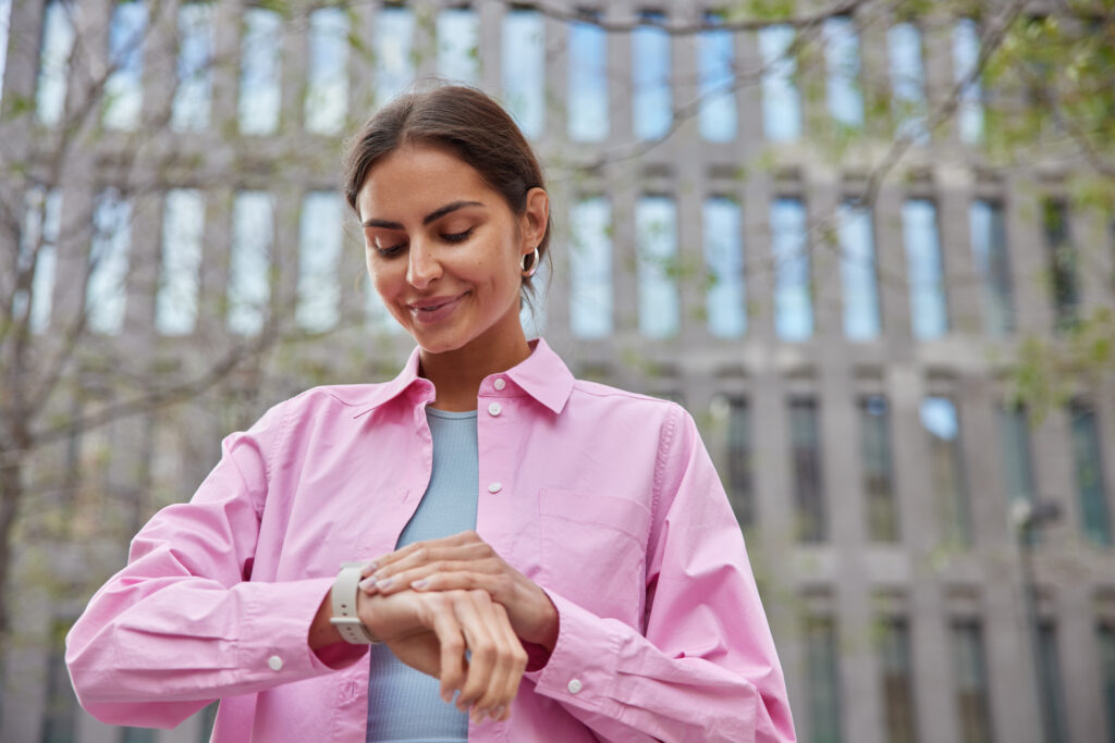 woman-looks-wrist-watch-checks-time-poses-outdoors-against-city-building