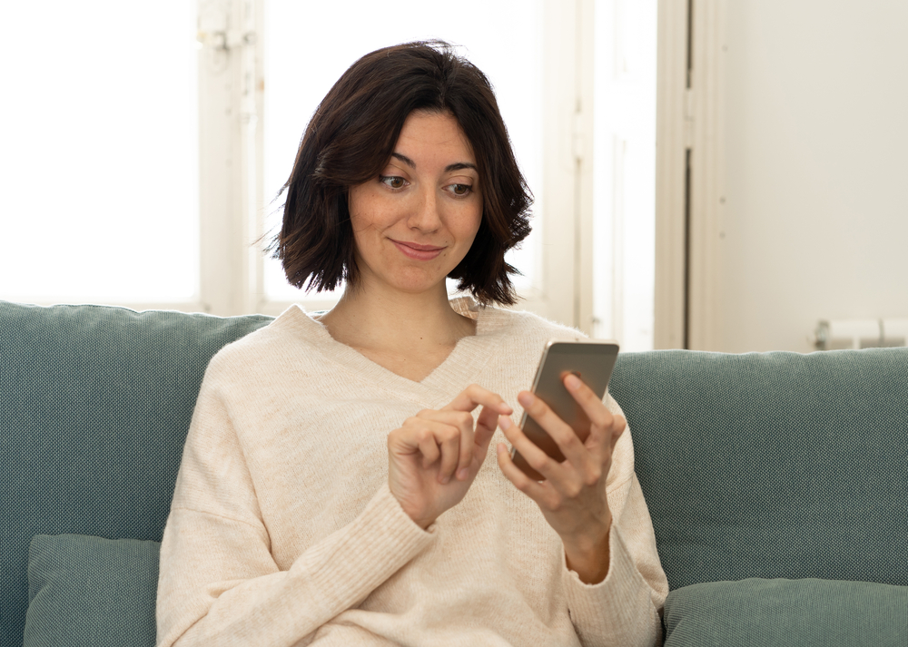 image-of-woman-sitting-on-couch-smirking-while-learning-dutch-via-an-application-on-her-smartphone