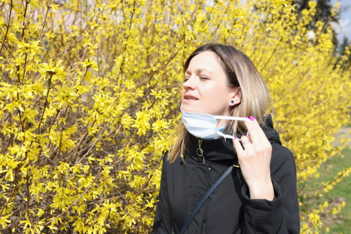 woman-with-closed-eyes-taking-off-face-mask-near-blossoming-tree