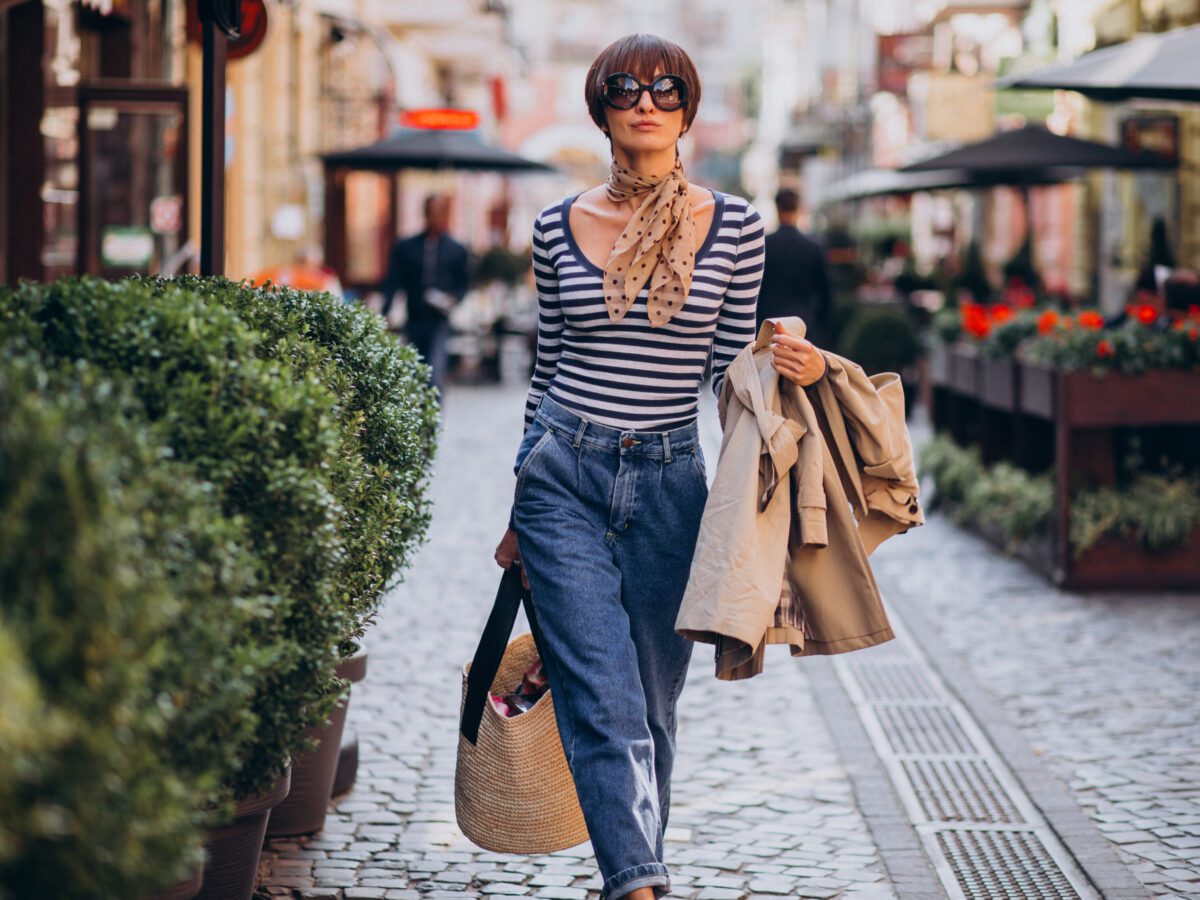 young-woman-in-sunglasses-with-striped-shirt-walking-down-street-surrounded-by-bushes