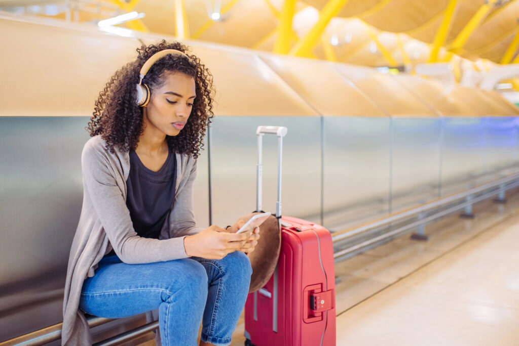 photo-of-young-woman-on-her-phone-with-headphones-waiting-in-airport-with-suitcase